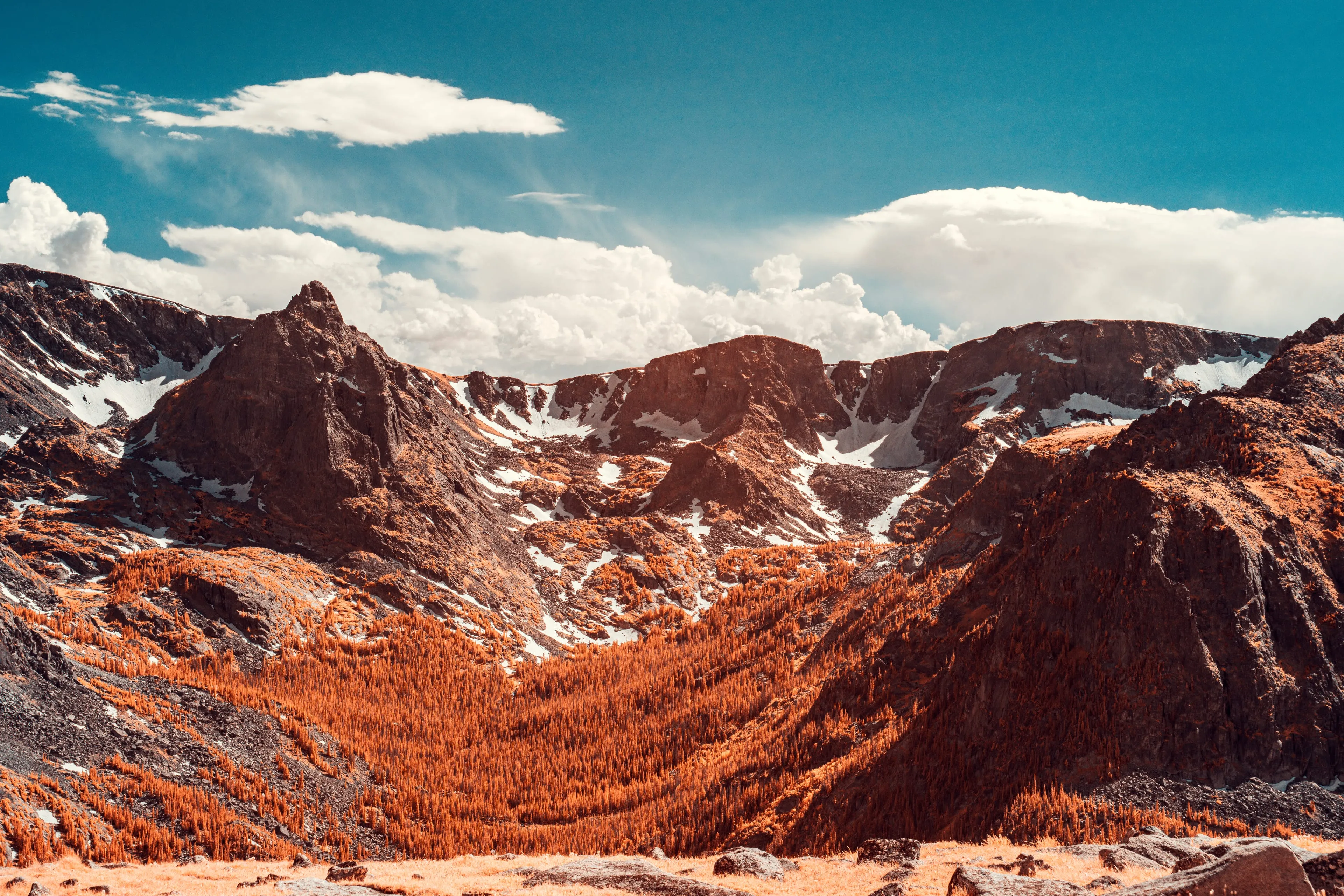 A high mountain cirque whose tundra floor is turned gold by infrared, its dark rock walls streaked with snow beneath massing white clouds.