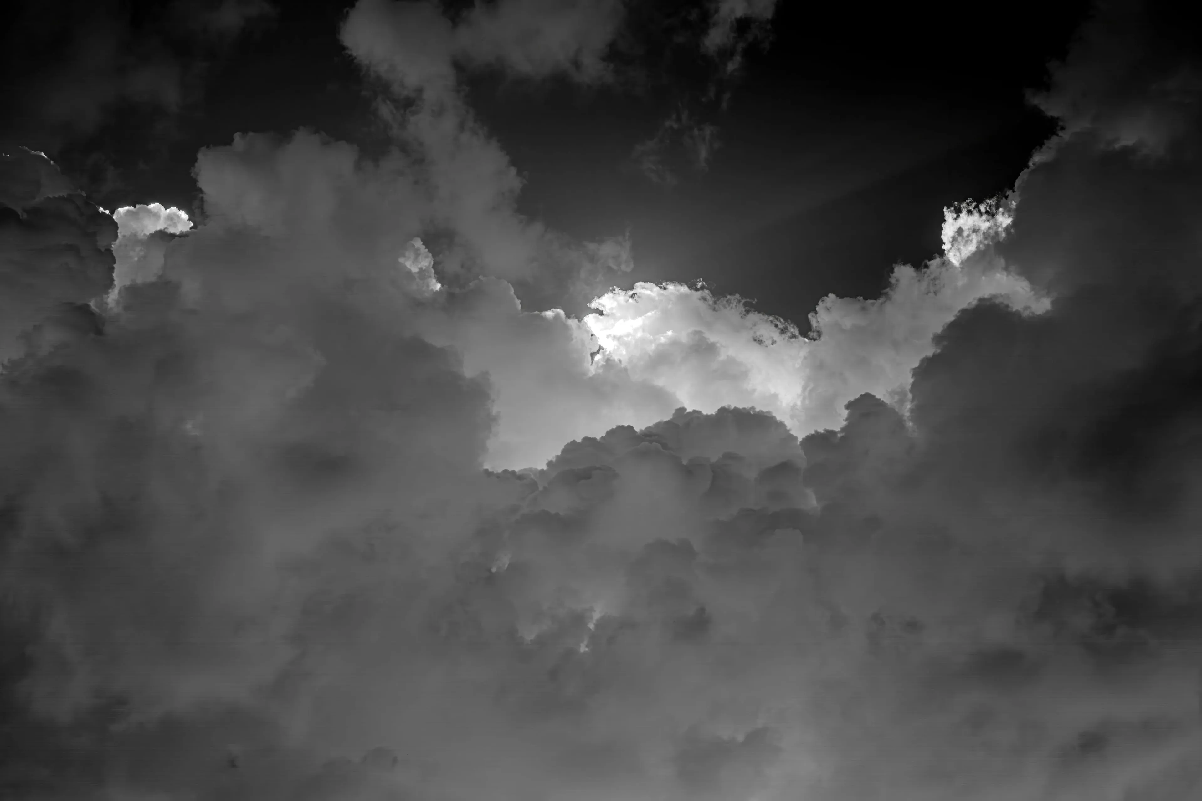 A black-and-white study of towering storm clouds, sunlight breaking through a dark sky above their billowing crowns.