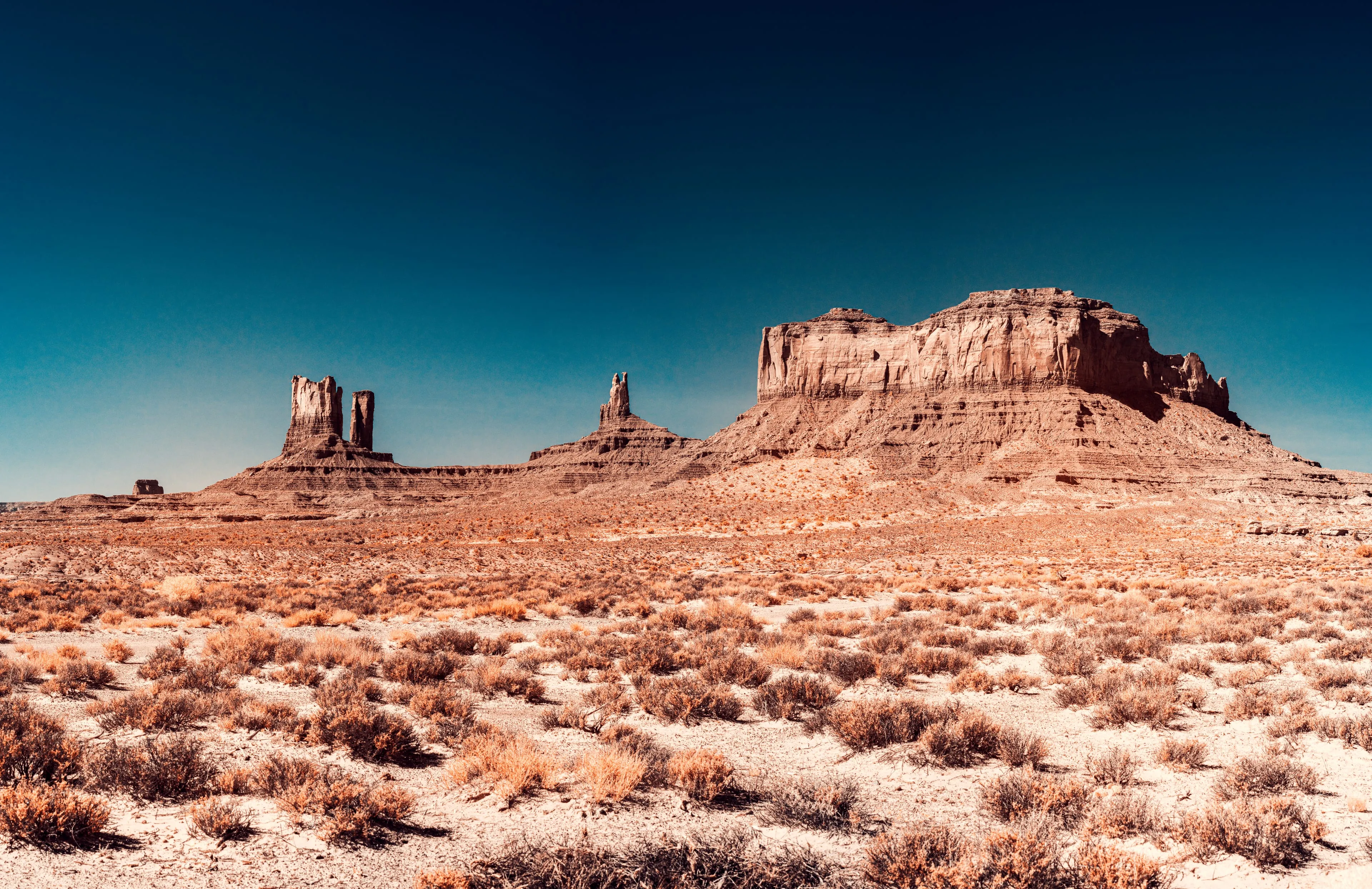 Sandstone buttes stand gathered on the desert floor of Monument Valley beneath a darkened sky, the ground scrub turned golden by infrared.