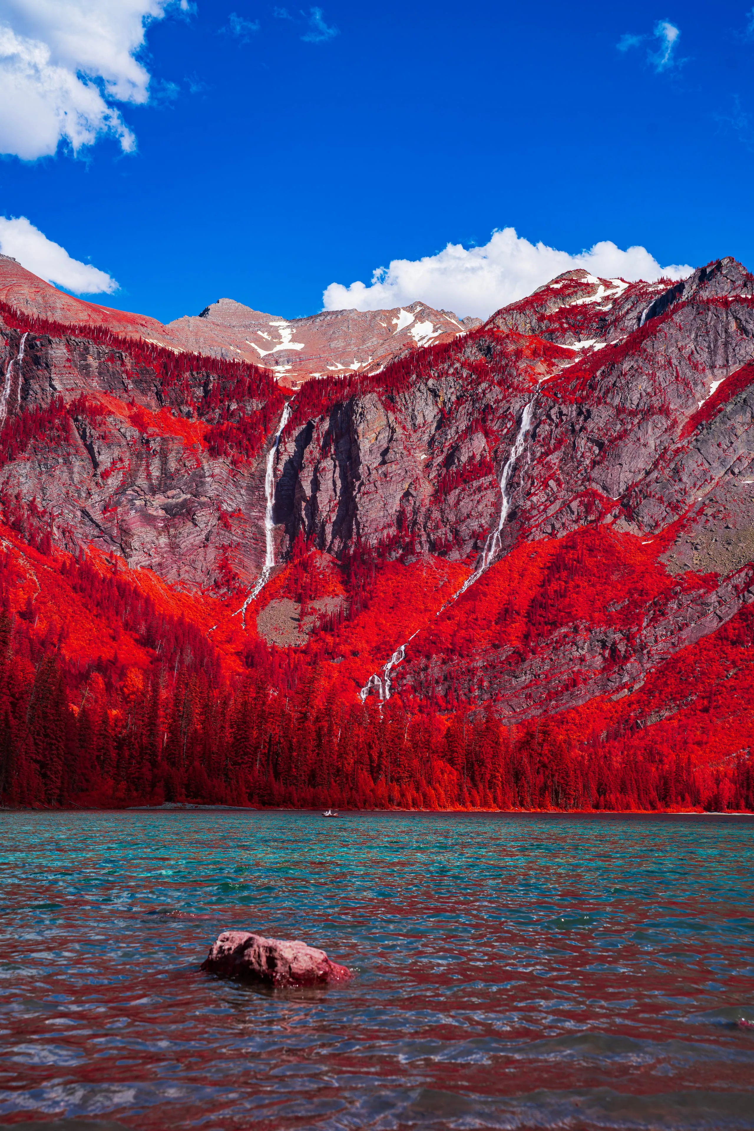 Thin white waterfalls descend a rugged mountain wall whose forest is turned red by infrared, toward a cold green lake.