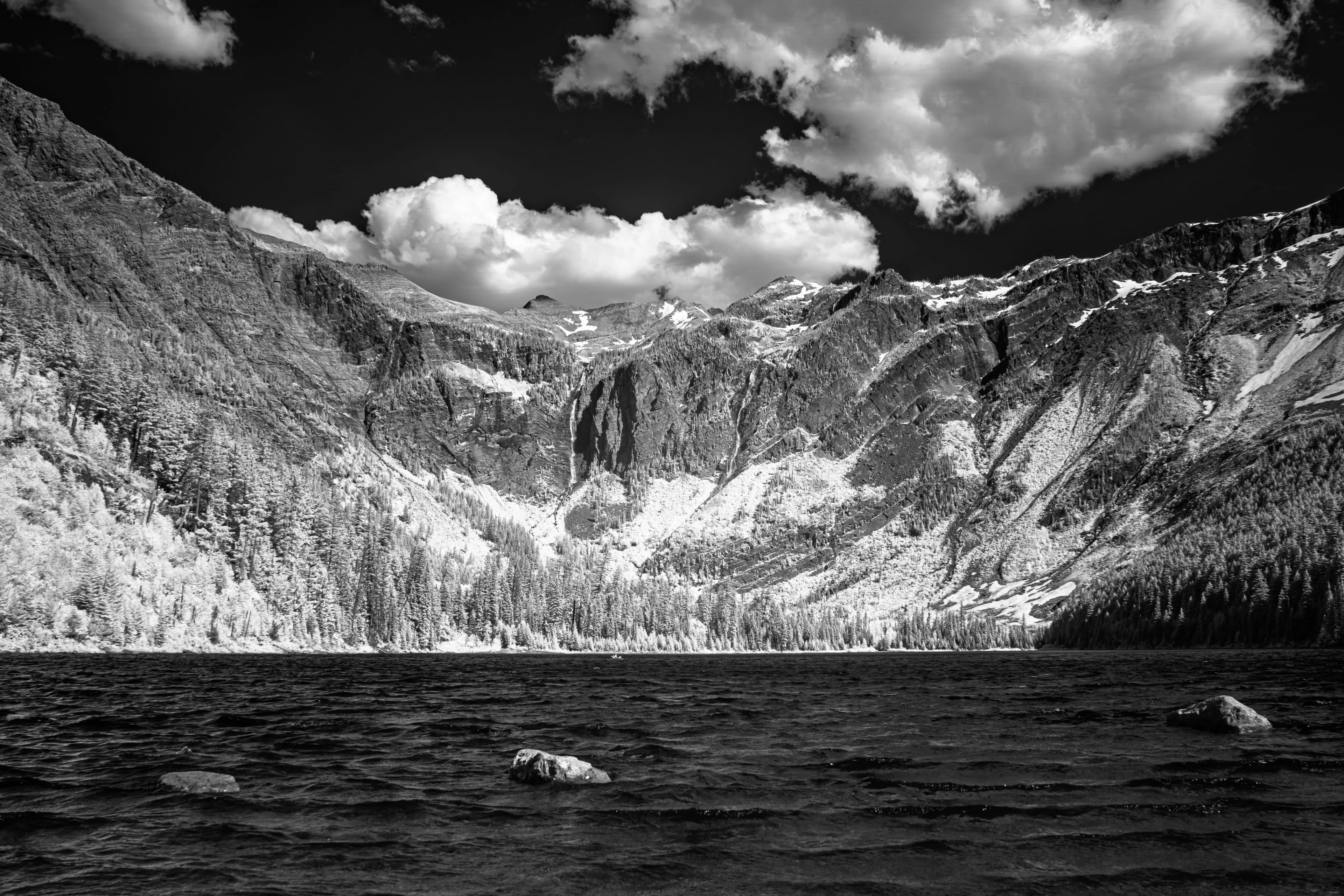 Infrared rendering of a Glacier National Park mountain wall whose forest burns white as bone beneath a pitch-black sky, thin pale waterfalls descending toward a dark lake where two bleached boulders break the surface.