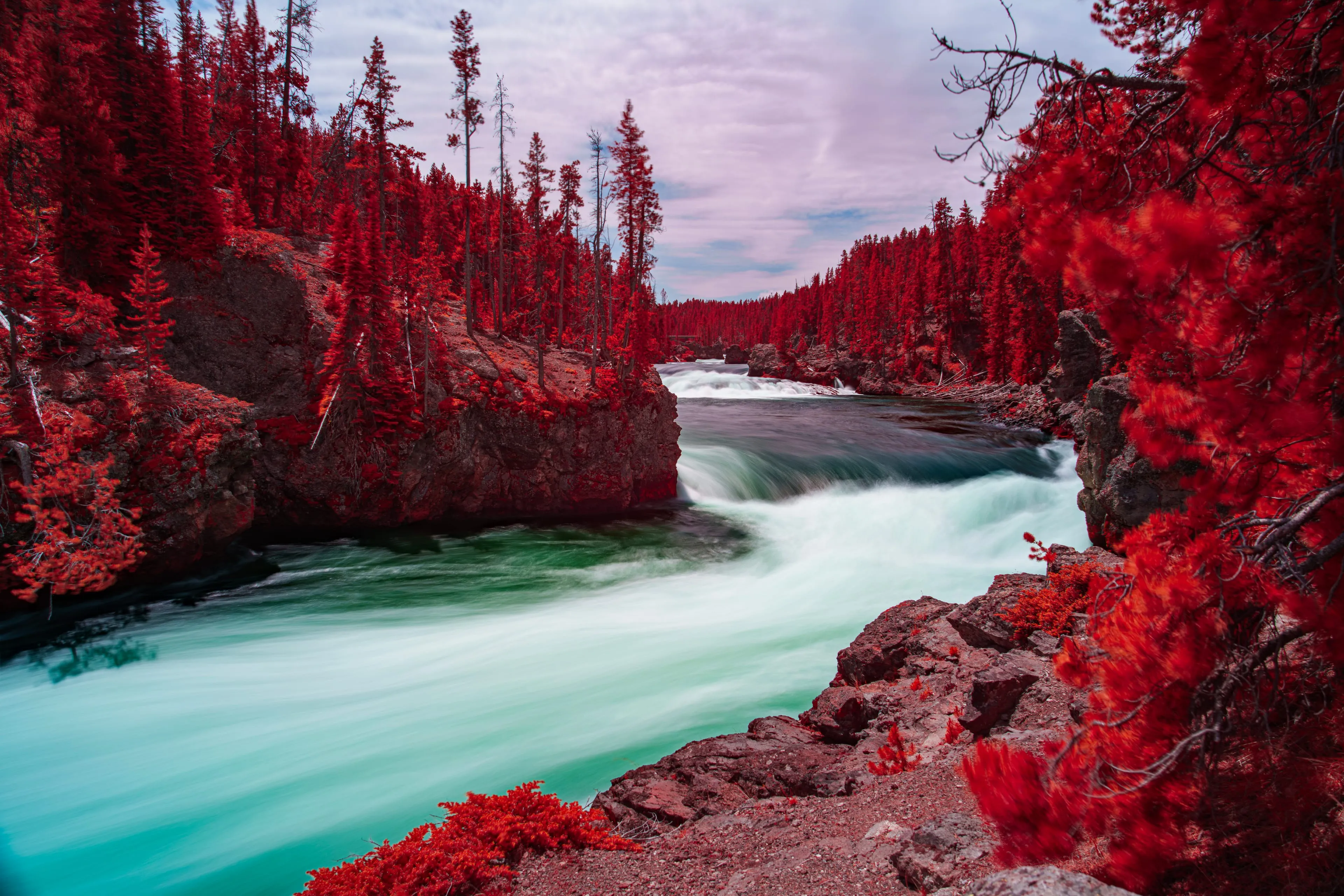 A long exposure smooths the river to silk as it races between banks turned red by infrared toward the brink of a waterfall at dusk.