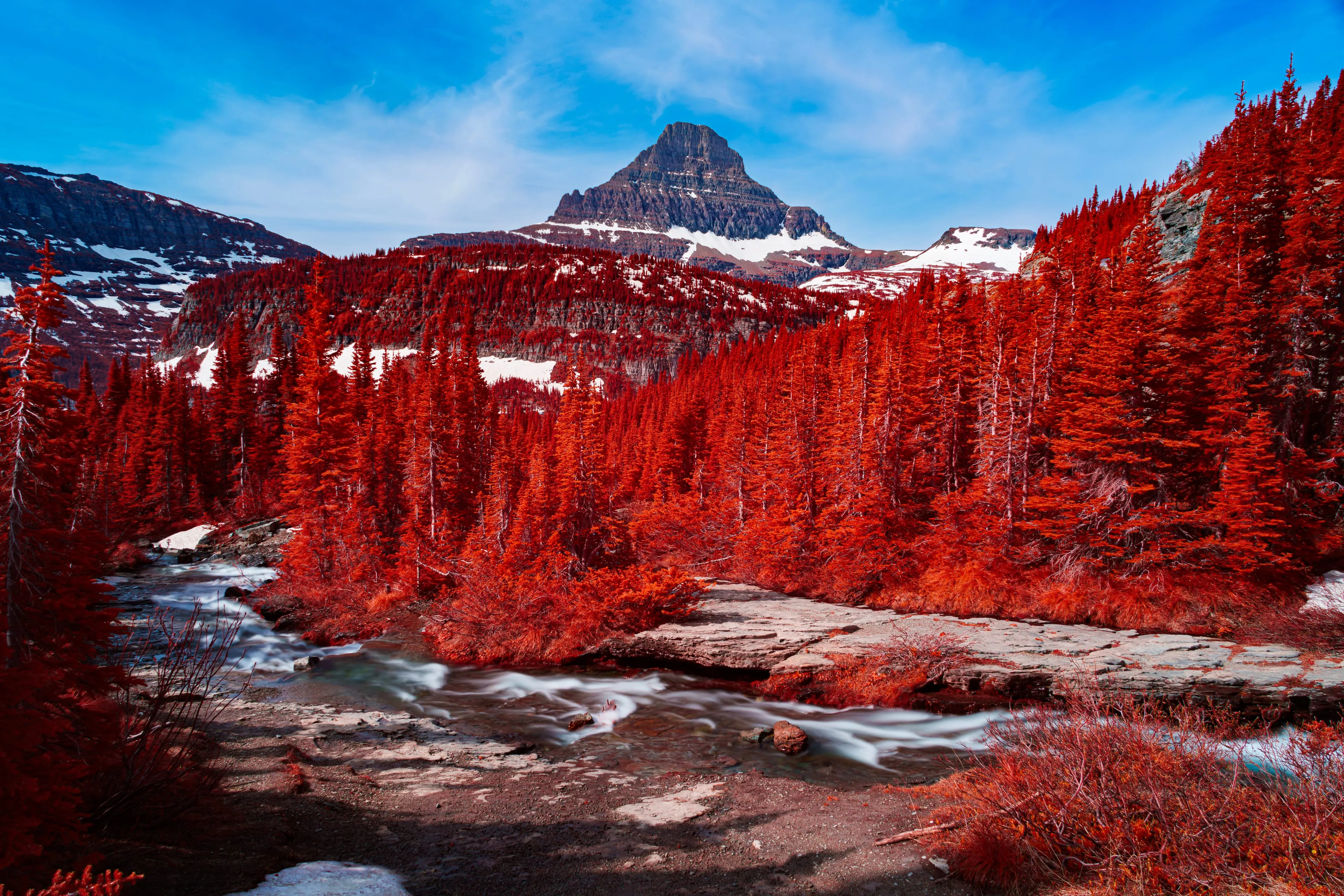 A bright creek rushes over rock ledges below a dark pyramidal peak, its banks crowded with forest turned red by infrared and patches of snow.