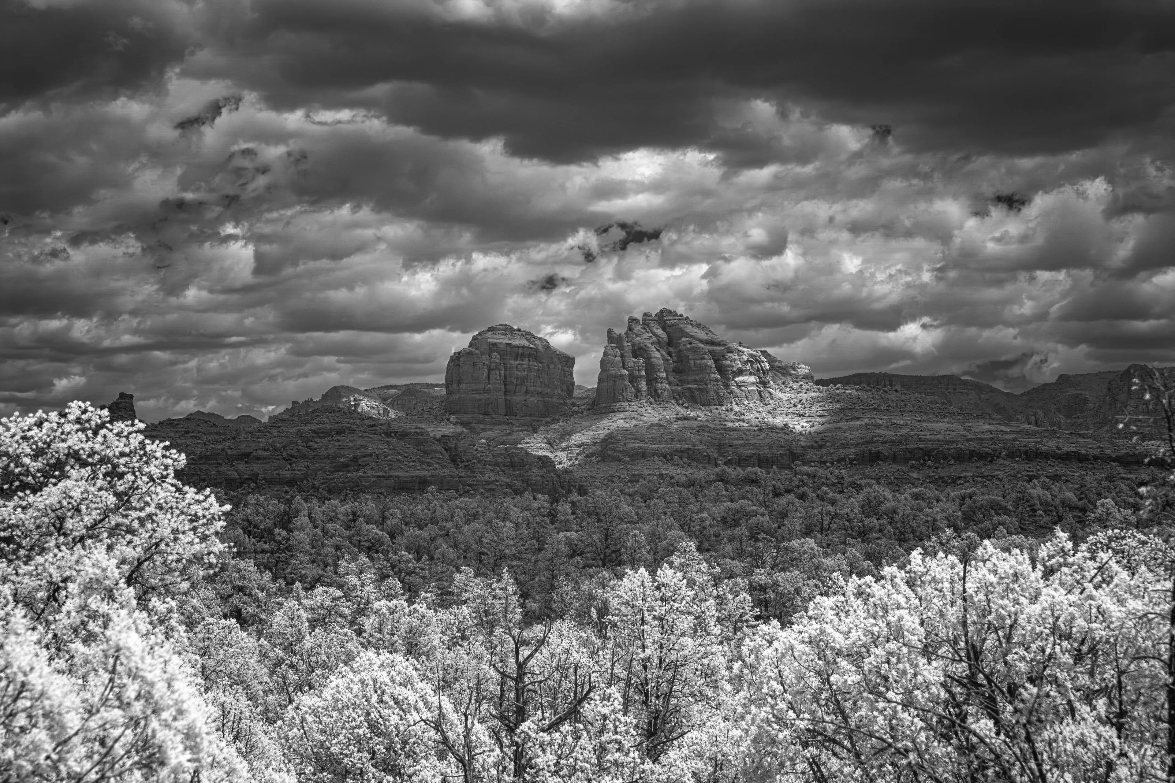A black-and-white infrared view of Sedona's sandstone buttes beneath brooding storm clouds, with pale glowing treetops in the foreground.
