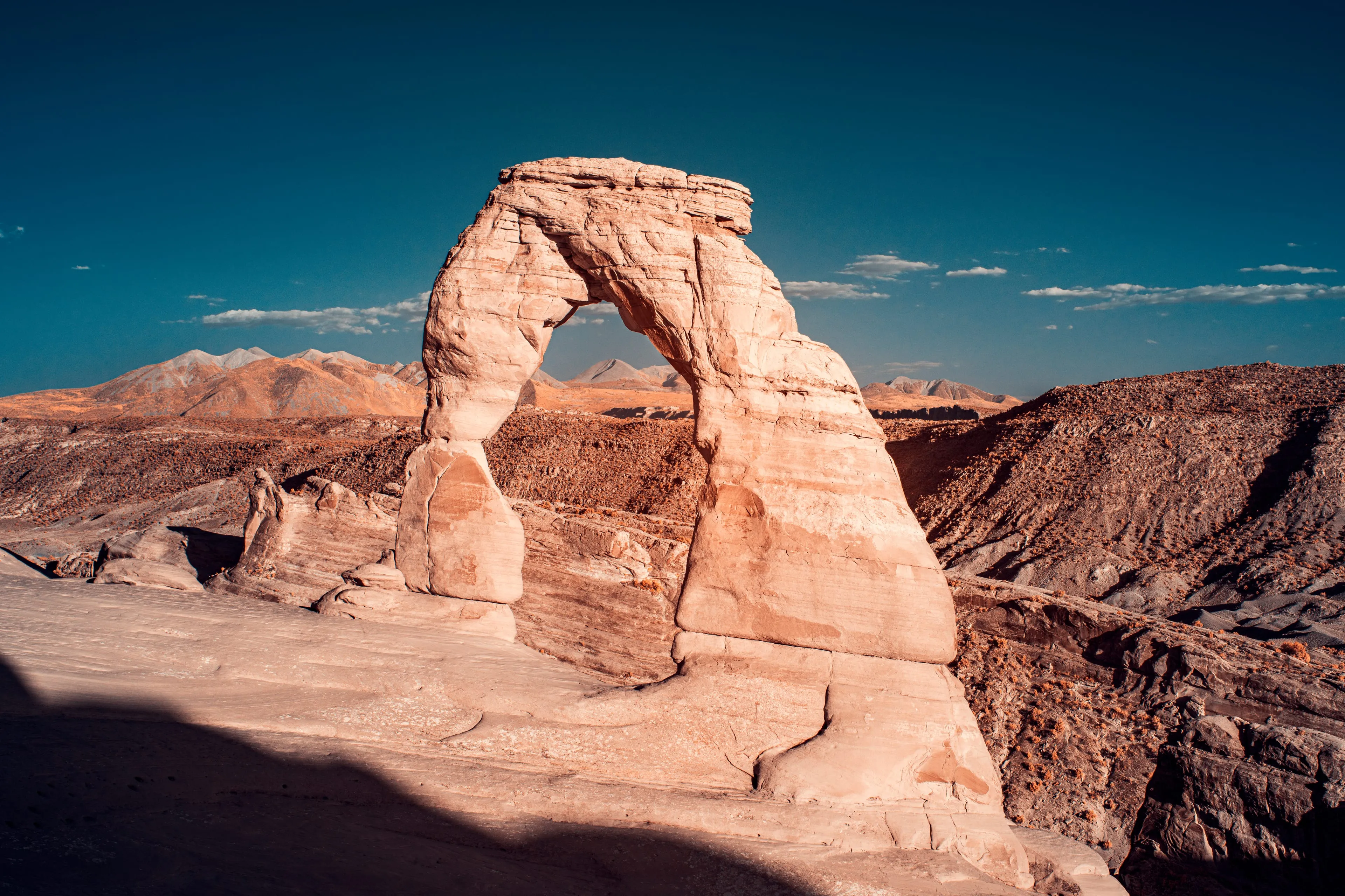 Delicate Arch stands gilded by low sun at the edge of a sandstone bowl, pale mountains visible through the opening.