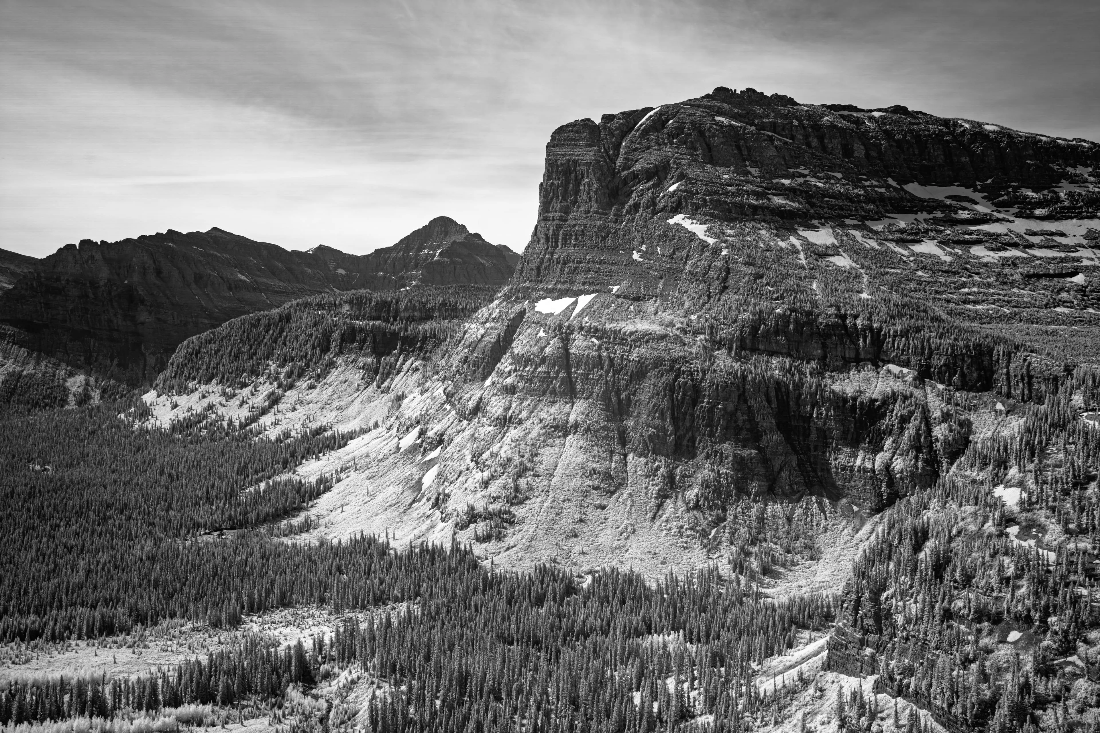 An infrared rendering of Heavy Runner Mountain in Glacier National Park, its dark layered stone standing imposing above a pale valley, with ranked peaks behind and a high faint sky.