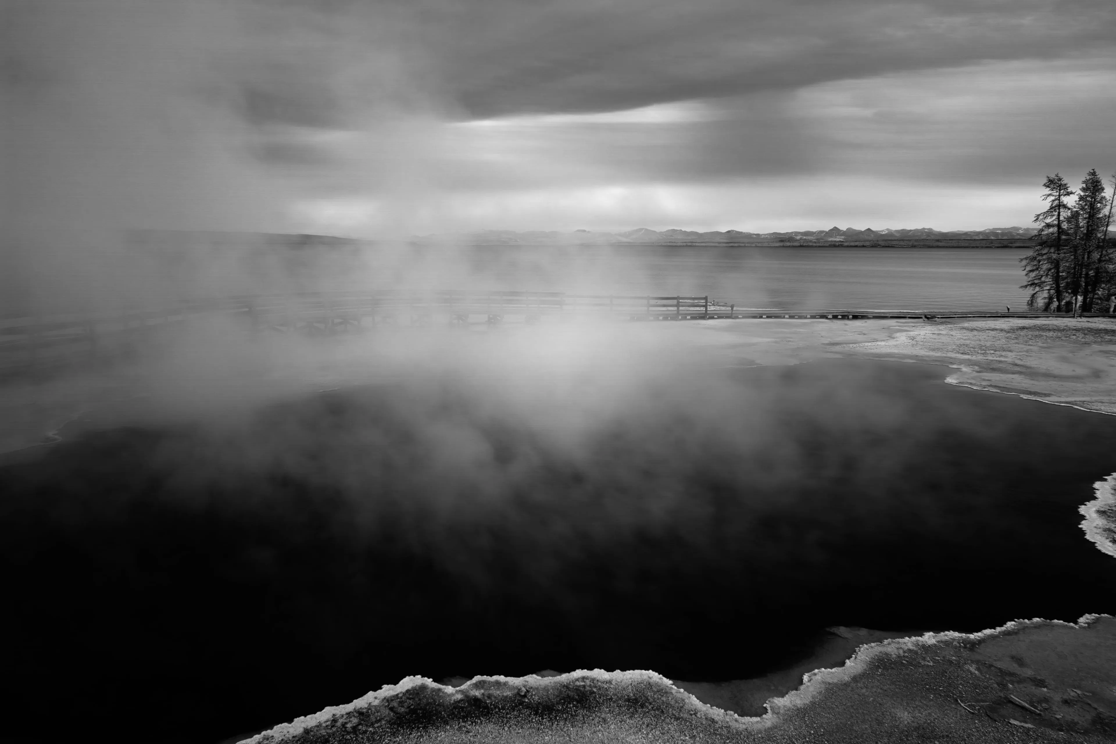 An infrared photograph of a Yellowstone hot spring rendered as a depthless black pool, with white steam rising off its surface and a pale crusted rim in the foreground, a dark lake and faint mountains beyond.