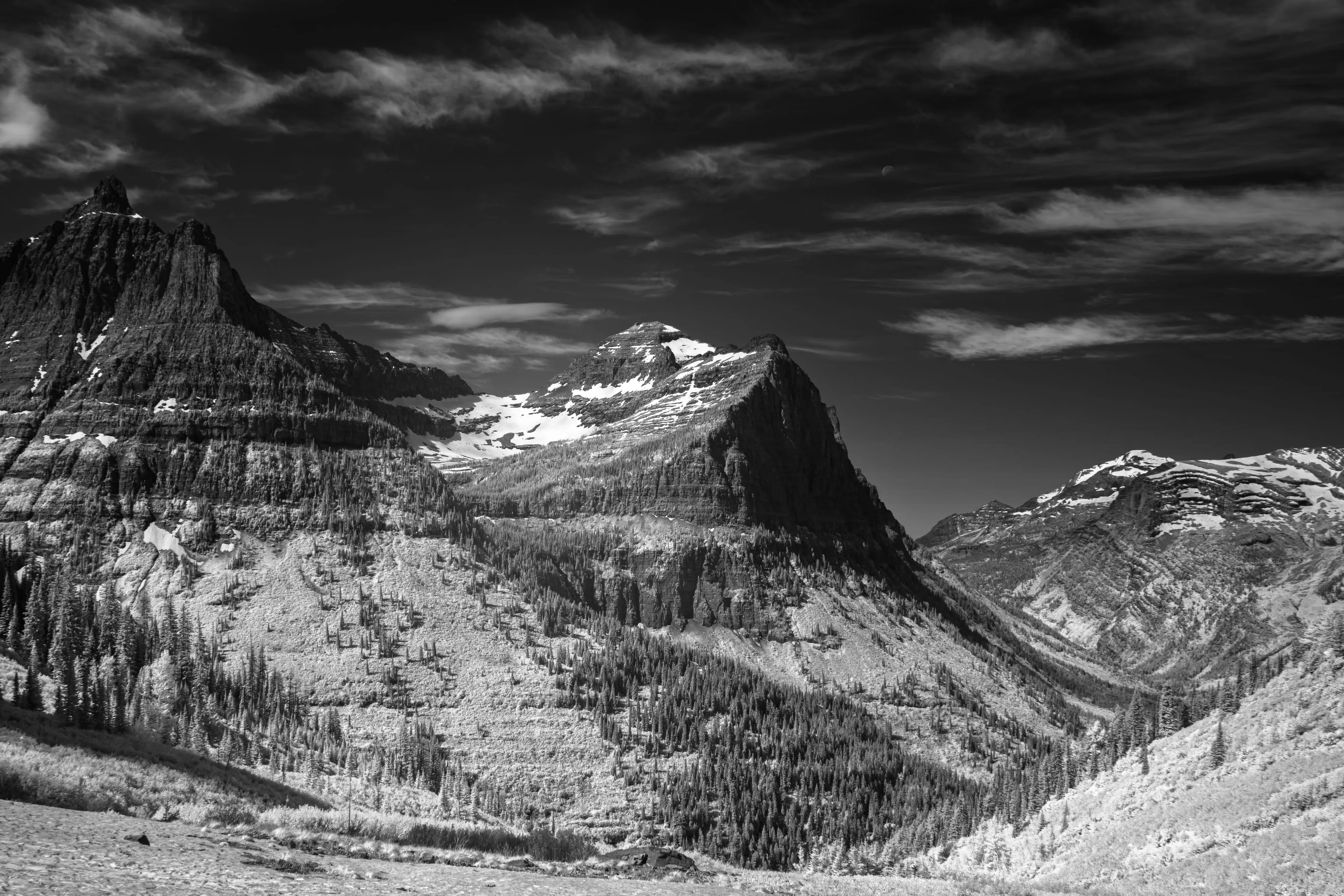 An infrared photograph of Glacier National Park: bone-white mountains and ridges beneath a near-black sky, with the small bright disc of the moon hanging above the central peak.