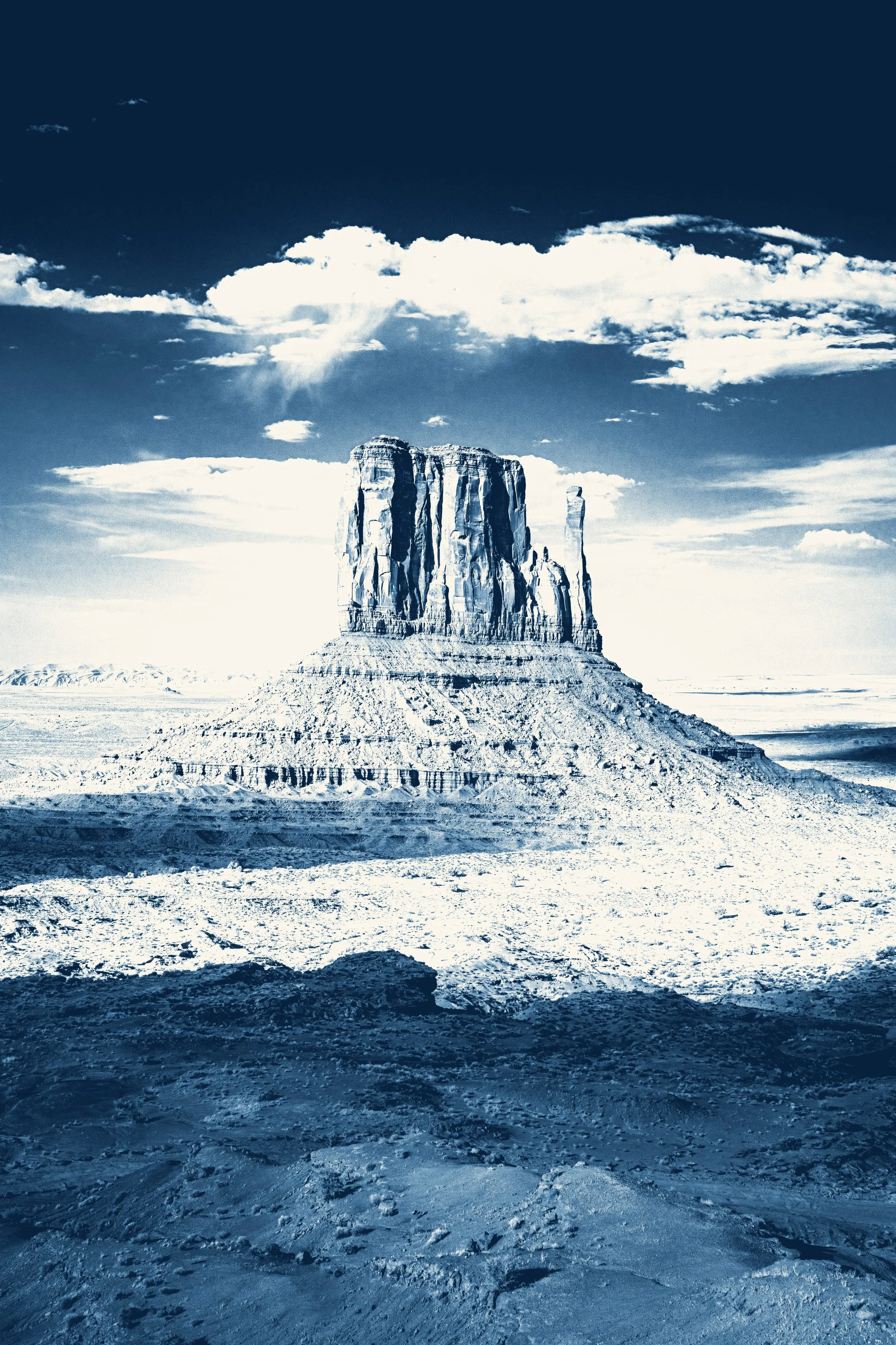 A lone sandstone butte rises from the desert floor of Monument Valley beneath massed white clouds, rendered in cyanotype blue.