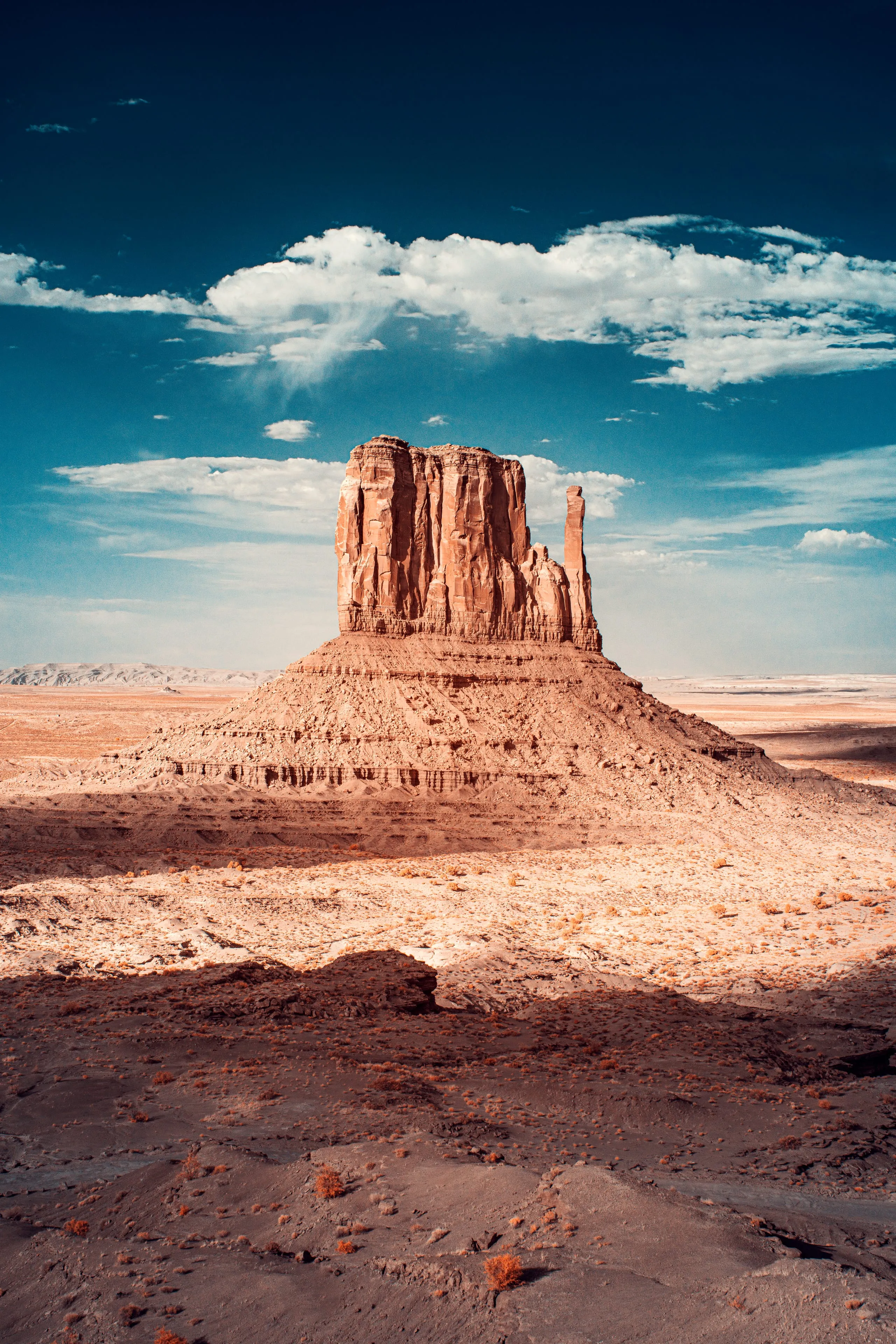 A lone sandstone mitten butte rises from the desert floor of Monument Valley beneath a deep blue sky and a single white cloud.