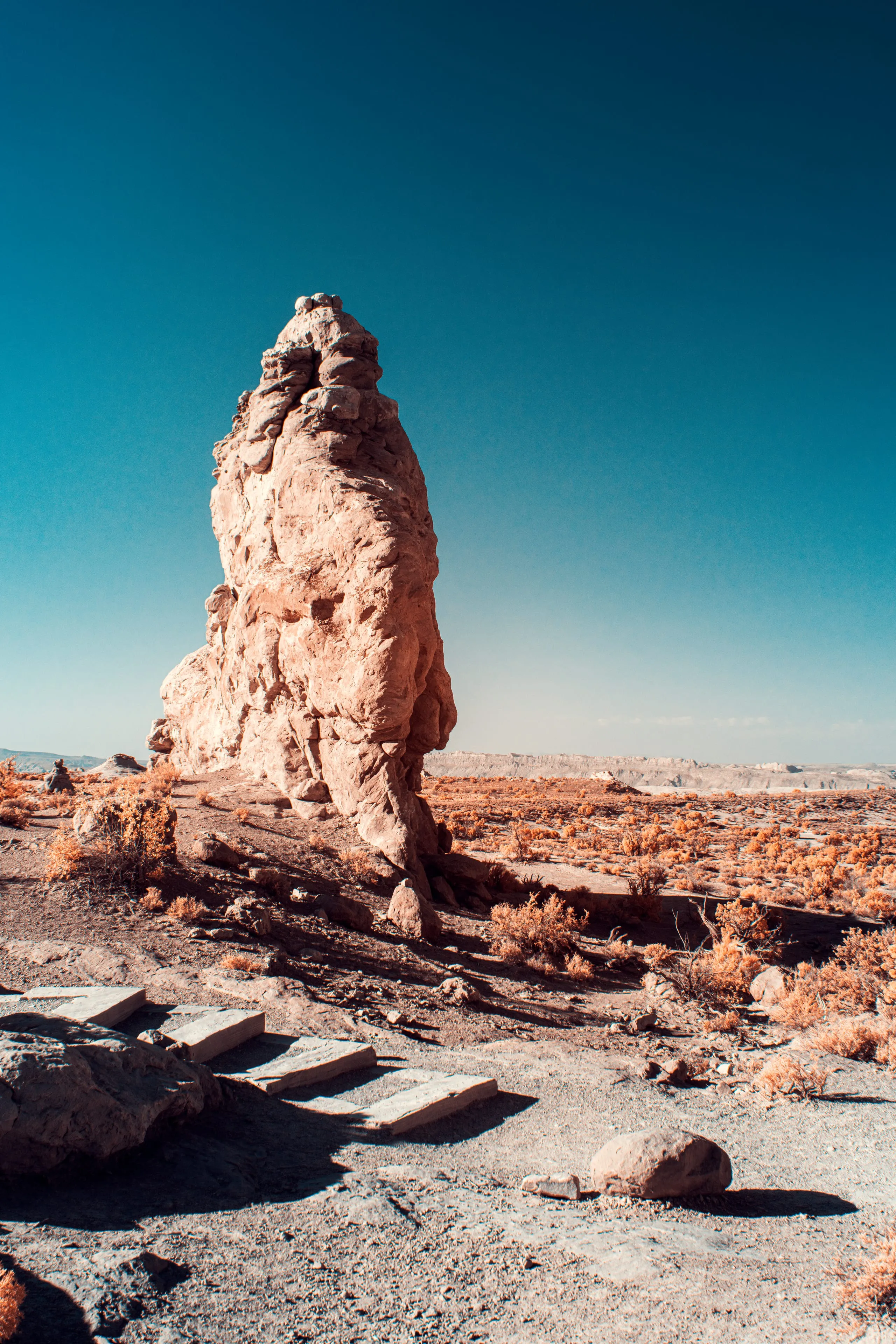 An infrared rendering of a single tall pinnacle of pale rock standing upright on a flat desert plain, lit hard beneath a deep blue sky, with rust-colored scrub and a few stone steps before it.