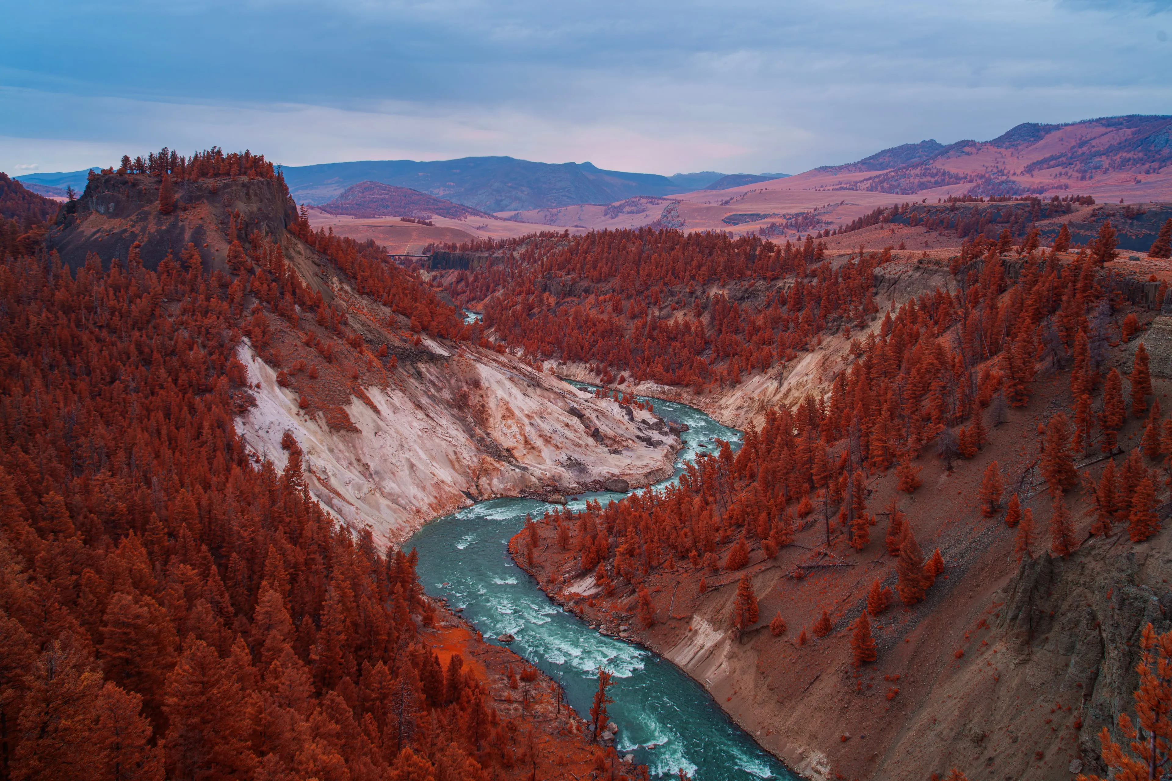 A turquoise river winds through a deep canyon at dusk, its forested slopes rendered blood-red by infrared beneath mauve hills and a pale pink sky.